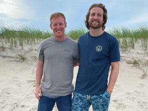 Zach McHale and Howie Proetto stand on the beach wearing blue and gray Dunebird t-shirts. The sun is shining in the background with a blue sky.
