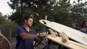 Surfer puts surfboard in the bed of a pickup truck at the entrance to the beach. He is wearing a blue terry towel pullover